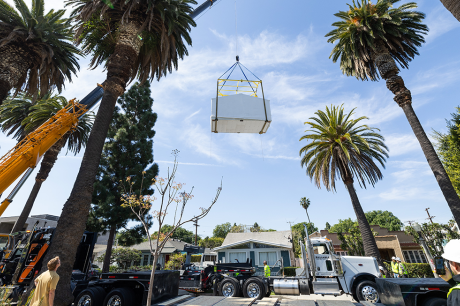 Crane lifting a large prefabricated structure over a residential street lined with tall palm trees and work trucks.