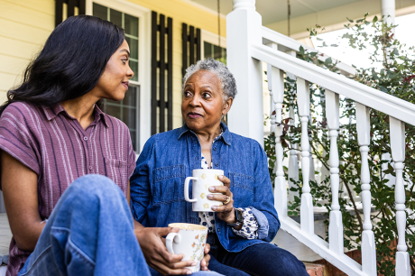 Image of an older woman of color and her daughter sitting on porch steps and drinking a warm beverage.  