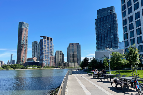 Waterfront walkway with benches and people, bordered by modern high-rise buildings along a river under a clear blue sky.