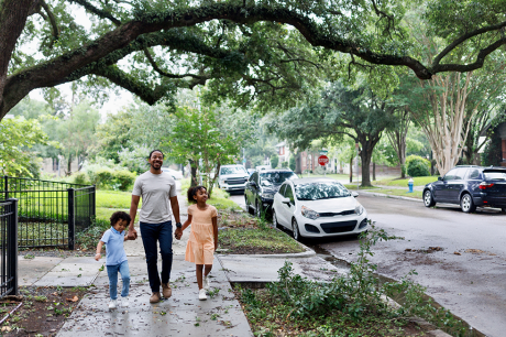 Image of a young Black father smiling and walking his kids down the street.