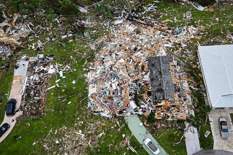 Aerial view of a neighborhood showing severe storm damage, with one home reduced to debris and wreckage scattered across surrounding yards.