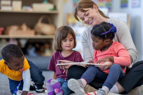 An adult sits with three young children on a classroom floor, reading a book together during a group learning activity.