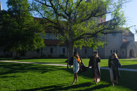 Three graduates in caps and gowns walk across a sunlit lawn in front of a large stone campus building and leafy trees.