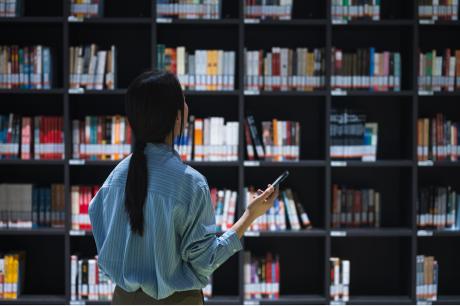 Person standing in front of tall library bookshelves, holding a smartphone while browsing rows of books.