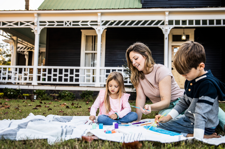 Photo of a mother and two young kids painting on the grass in front of their rural home. 