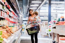 A mother with an adorable baby on her hip carries a shopping basket through the produce aisle in a grocery store.