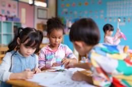 Three young children sit at a table, coloring, painting, and drawing in a colorful classroom.