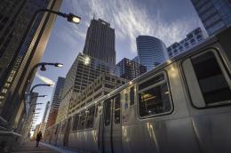 A close upward angle of a silver public train against a backdrop of skyscrapers and a whispy sky.