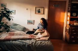A young woman with long, curly hair and a long-sleeve blue shirt sits on a low bed in her apartment, petting an adorable cat and holding a mug of something in her other hand.
