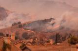 Smoke billows and a wildfire blazes on a hilltop adjacent to a neighborhood in an arid, hilly region.