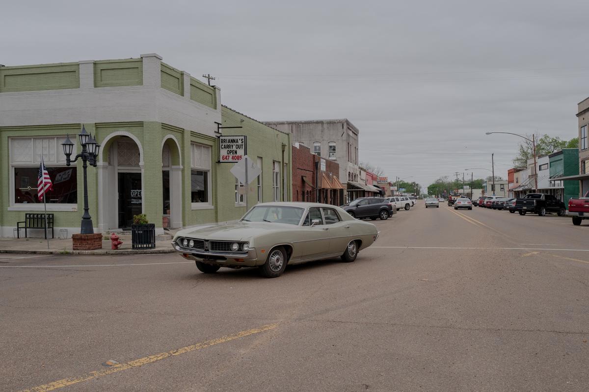 An older, green car turning down a commercial street. 